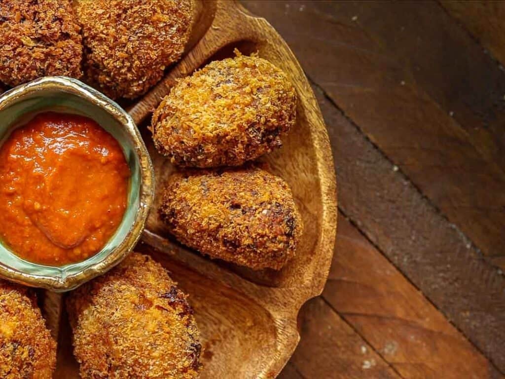 A wooden plate with crispy, breaded kimchi fried rice balls arranged around a small bowl of red dipping sauce, set on a rustic wooden table.