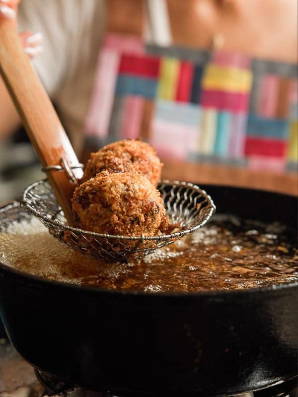 A person uses a slotted fryer spoon to lift two golden-brown, breaded balls from hot oil in a black pan. The background shows someone wearing a colorful, patterned apron.