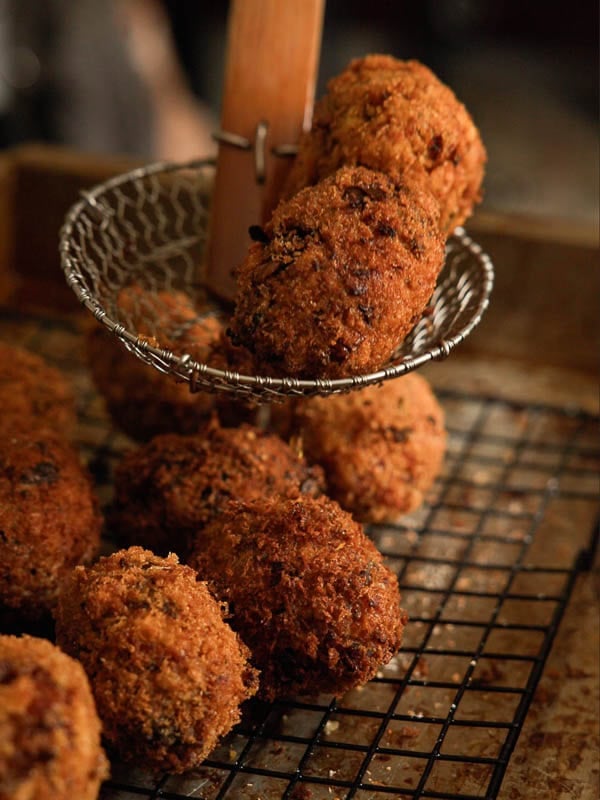 A wire skimmer lifts two golden-brown, crispy croquettes from a cooling rack holding several more croquettes on a baking tray.