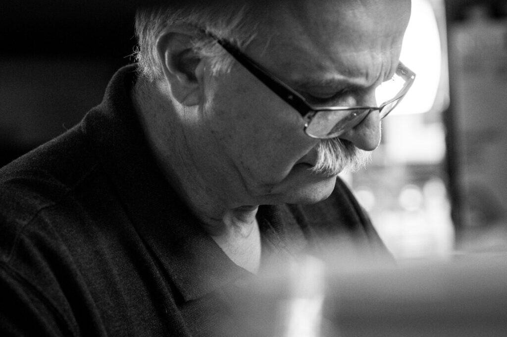 Black and white close-up of an older man with glasses and a mustache, looking down with a focused expression, wearing a dark collared shirt. The background is blurred.