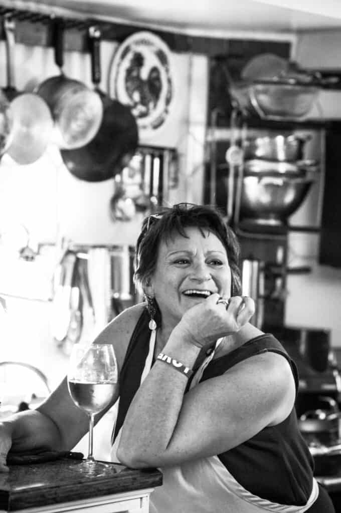 A woman wearing an apron sits at a kitchen counter, smiling and laughing with her chin resting on her hand. A glass of white wine is in front of her, and pots and kitchenware hang in the background. The photo is in black and white.
