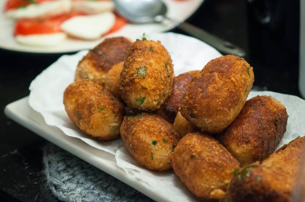 A plate of golden-brown croquettes sits on a white napkin, garnished with herbs. In the background, a spoon and a dish with sliced tomatoes and mozzarella are visible.