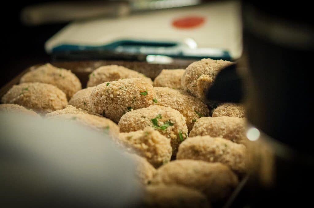 A close-up view of a tray filled with breaded, oval-shaped croquettes garnished with bits of green herbs, ready to be cooked. The background is softly blurred.