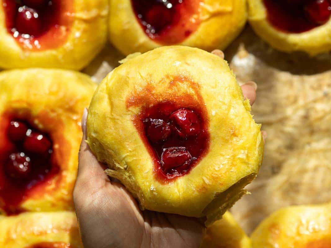 Easy Cherry Brioche Sweet Roll - Vegan Pastry. 19 A hand holds a cherry brioche sweet roll, surrounded by more similar pastries on a baking sheet.