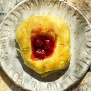 Easy Cherry Brioche Sweet Roll - Vegan Pastry. 36 A round cherry brioche sweet roll with a golden, fluffy crust and a center filled with red cherry filling sits on a textured ceramic plate.