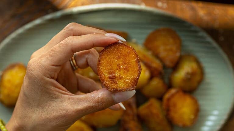 A hand with neatly manicured nails holds up a crispy, golden-brown roasted potato slice as part of a roasted potatoes recipe. More roasted potato slices are visible on a plate in the background.