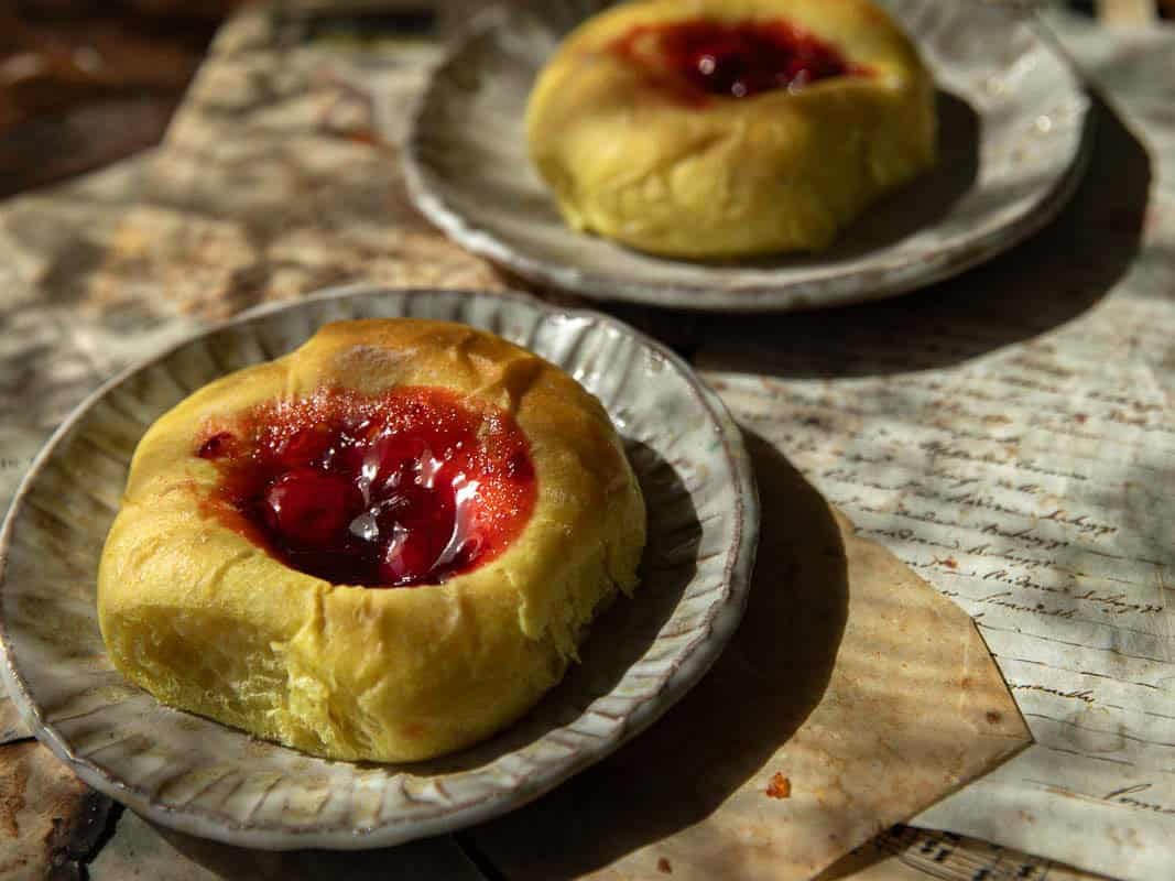Easy Cherry Brioche Sweet Roll - Vegan Pastry. 20 Two round cherry brioche sweet rolls are placed on ceramic plates, set atop sheets of handwritten paper in warm, natural sunlight.