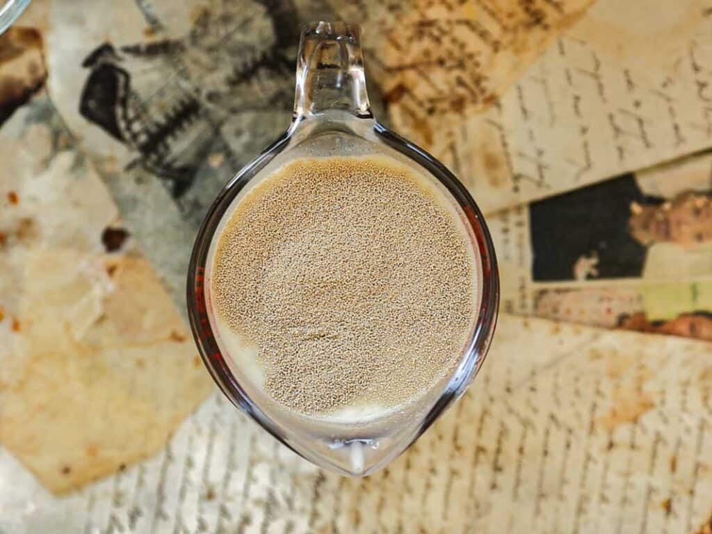 Easy Cherry Brioche Sweet Roll - Vegan Pastry. 21 A close-up of an overhead glass measuring cup filled with yeast dissolving in water, placed on top of scattered vintage papers and handwritten letters.