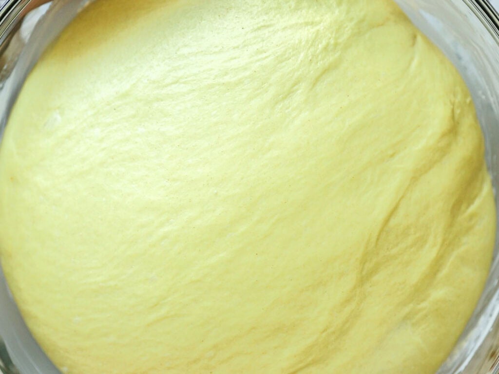 Easy Cherry Brioche Sweet Roll - Vegan Pastry. 26 Close-up of smooth, yellow bread dough rising in a glass bowl, showing a soft and airy texture on the surface.
