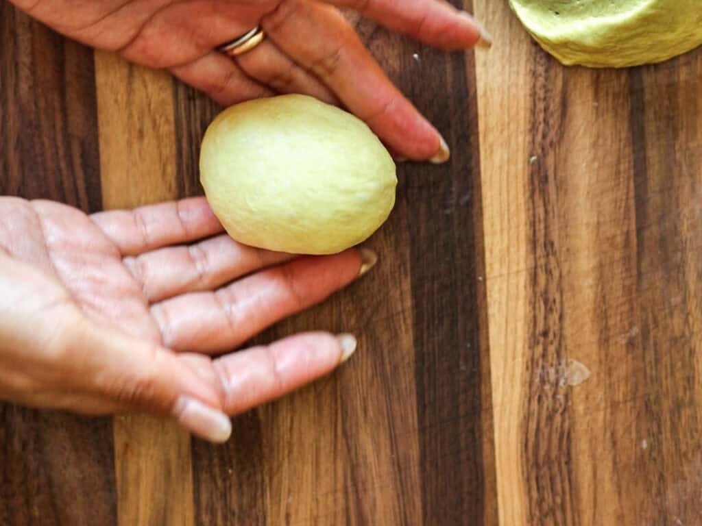 Easy Cherry Brioche Sweet Roll - Vegan Pastry. 28 Two hands shape a piece of dough into an oval on a wooden surface, with another dough ball visible nearby.