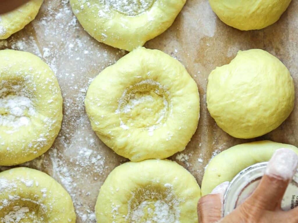 Easy Cherry Brioche Sweet Roll - Vegan Pastry. 30 Close-up of several dough balls on a floured parchment surface, some with indents in the center. A hand is sprinkling flour from a small container onto the dough.