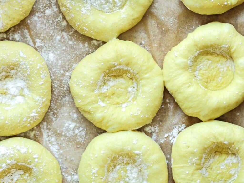 Easy Cherry Brioche Sweet Roll - Vegan Pastry. 31 Unbaked dough rounds with indented centers are arranged on a floured surface, ready for baking. The dough appears soft and slightly yellow, dusted with flour.