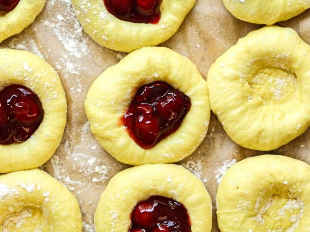 Easy Cherry Brioche Sweet Roll - Vegan Pastry. 32 Unbaked pastry dough rounds on parchment paper, some filled with cherry jam and others with empty indents, lightly dusted with flour.