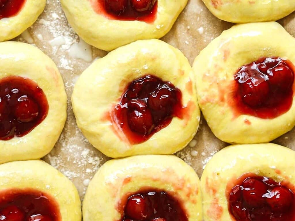 Easy Cherry Brioche Sweet Roll - Vegan Pastry. 33 Close-up of unbaked pastries with yellow dough, each topped with a dollop of cherry or berry fruit filling, arranged in rows on a floured surface.
