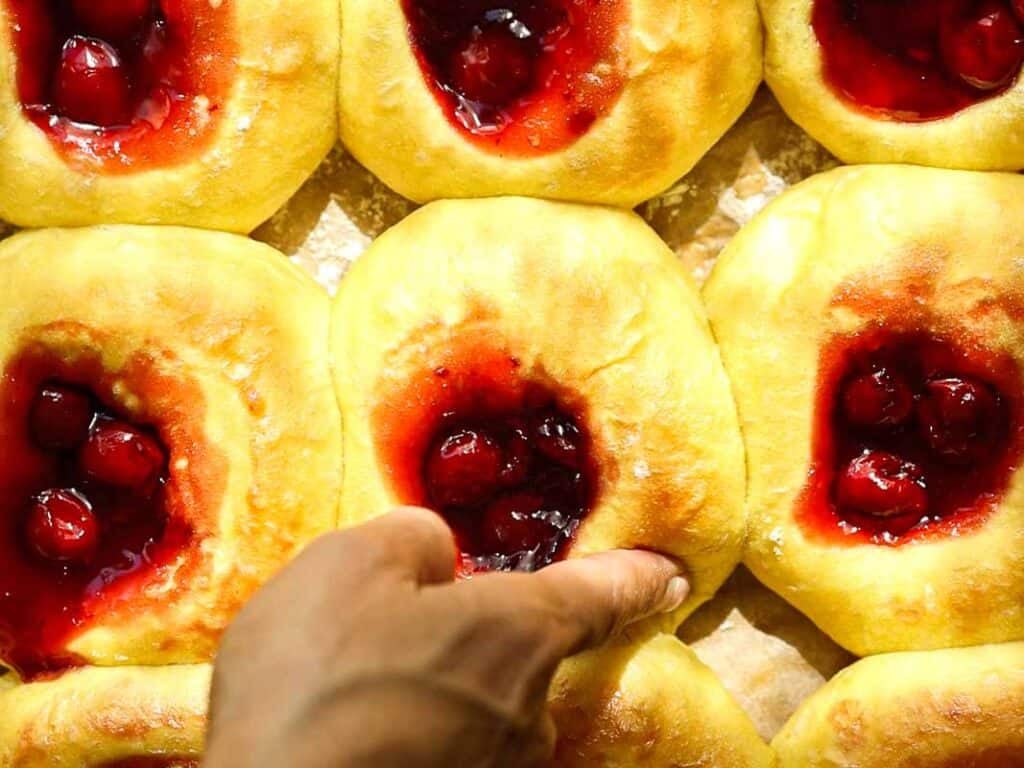 Easy Cherry Brioche Sweet Roll - Vegan Pastry. 35 A hand presses into a soft, round pastry topped with cherry filling, surrounded by several similar pastries on a baking sheet. The pastries are golden-brown and look freshly baked.
