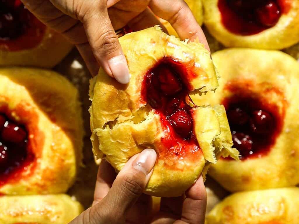 Easy Cherry Brioche Sweet Roll - Vegan Pastry. 18 Hands breaking apart a soft yellow cherry brioche sweet roll filled with bright red cherry filling, surrounded by similar pastries in the background.