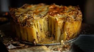 A close-up of a baked rigatoni pie with rigatoni standing upright, covered in melted cheese and sauce, with a wedge removed to reveal the inside. The pie sits on a rustic surface with some sauce spilled around.