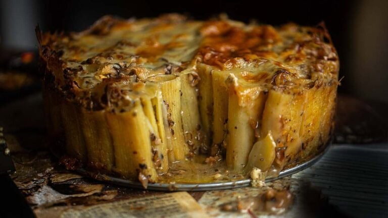 Recipes 1 A close-up of a baked rigatoni pie with rigatoni standing upright, covered in melted cheese and sauce, with a wedge removed to reveal the inside. The pie sits on a rustic surface with some sauce spilled around.