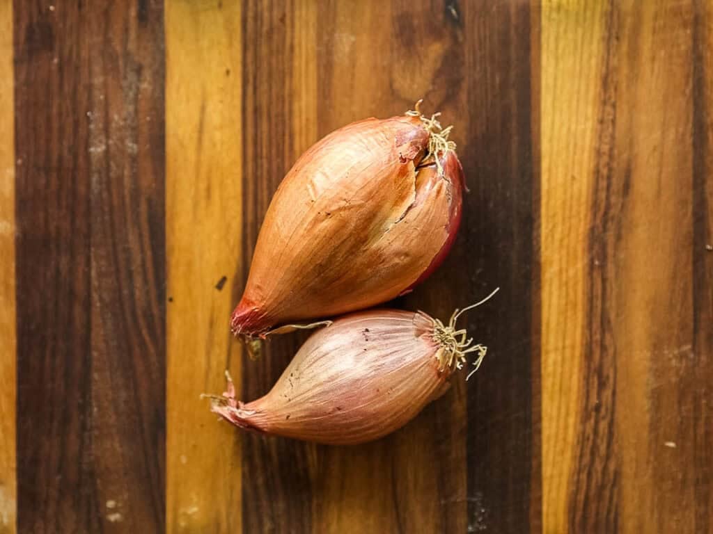Two whole shallots with papery skin and roots attached, resting on a striped wooden cutting board.