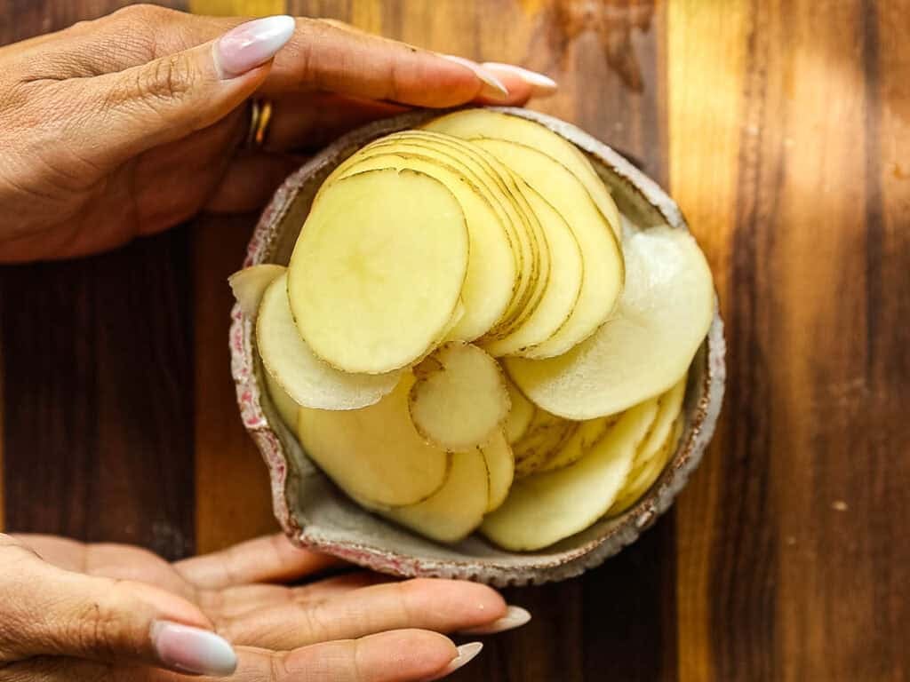 A bowl filled with thinly sliced raw potatoes is held gently by two hands over a wooden surface.