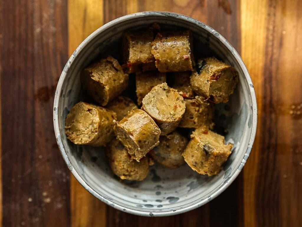 A ceramic bowl filled with several pieces of sliced vegan sausage, placed on a wooden surface with striped patterns.