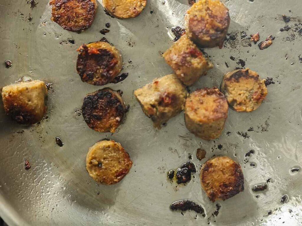 Several browned sausage slices cooking in a stainless steel pan, with some pieces showing dark seared edges and bits of oil and seasoning scattered around.