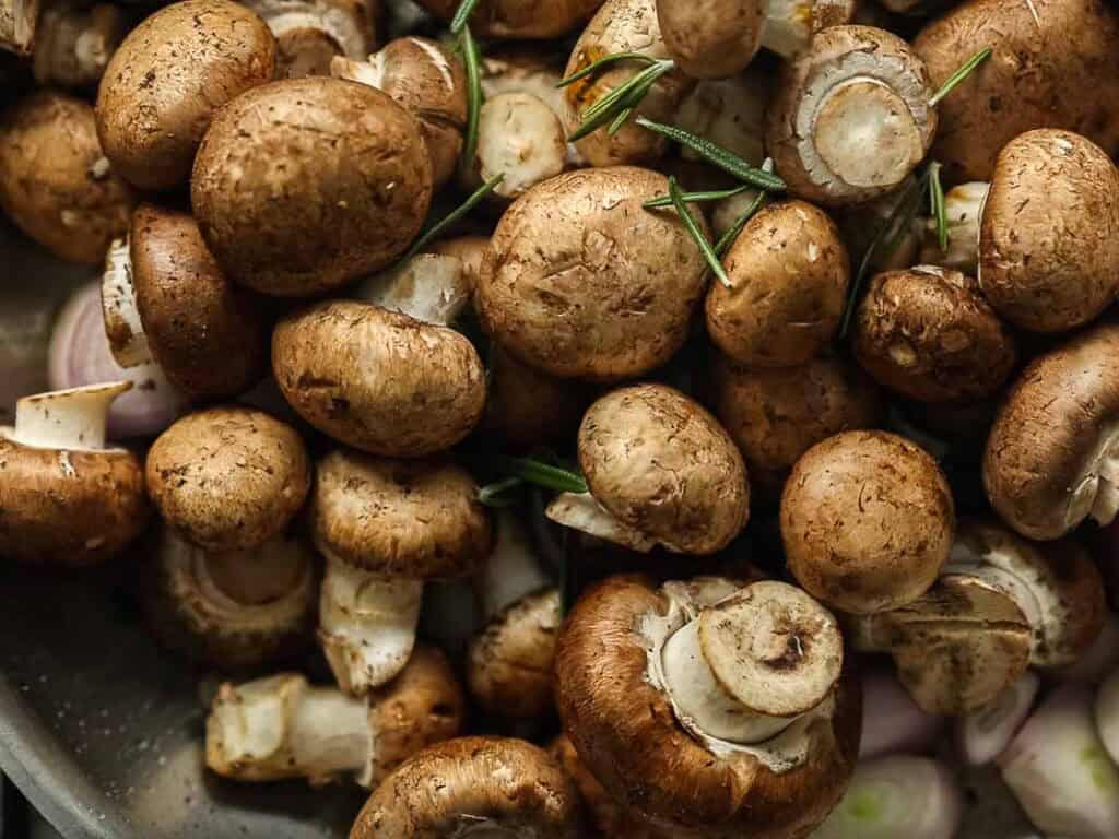 A close-up of fresh brown mushrooms with sprigs of rosemary scattered among them. The mushrooms have visible stems and some sliced caps, creating a rustic, earthy appearance.
