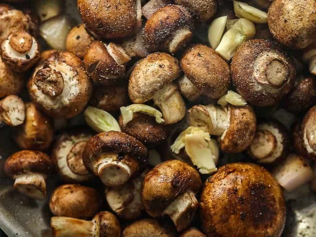 Whole cremini mushrooms and chunks of garlic cloves are being cooked together, with visible seasonings and a slight sheen from oil or butter on their surfaces.