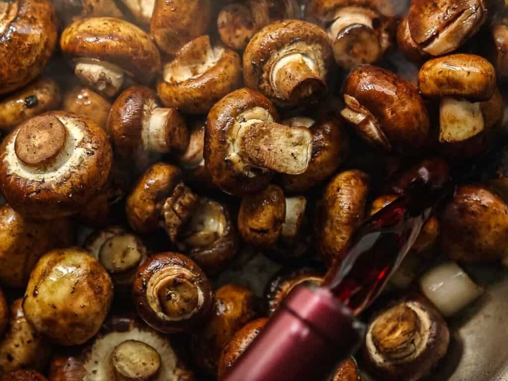 A close-up of brown mushrooms being cooked in a pan, with red wine being poured over them.