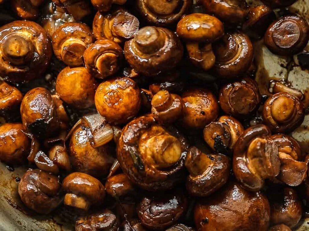 A close-up of sautéed brown mushrooms, glistening with a rich, glossy coating, showing their rounded caps and stems, cooked together in a pan.