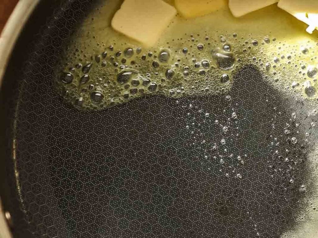 Close-up of butter melting in a pan, with small bubbles forming on the surface and several cubes of butter visible at the top edge of the image.