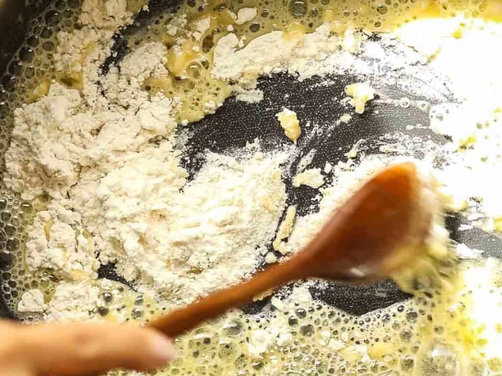 A wooden spoon stirs flour into melted butter in a pan, starting the process of making a roux.
