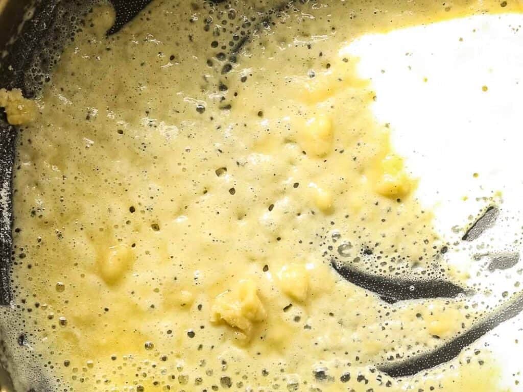 A close-up of melted butter and flour being whisked together in a pan, forming a bubbly and slightly lumpy roux, commonly used as a base for sauces.