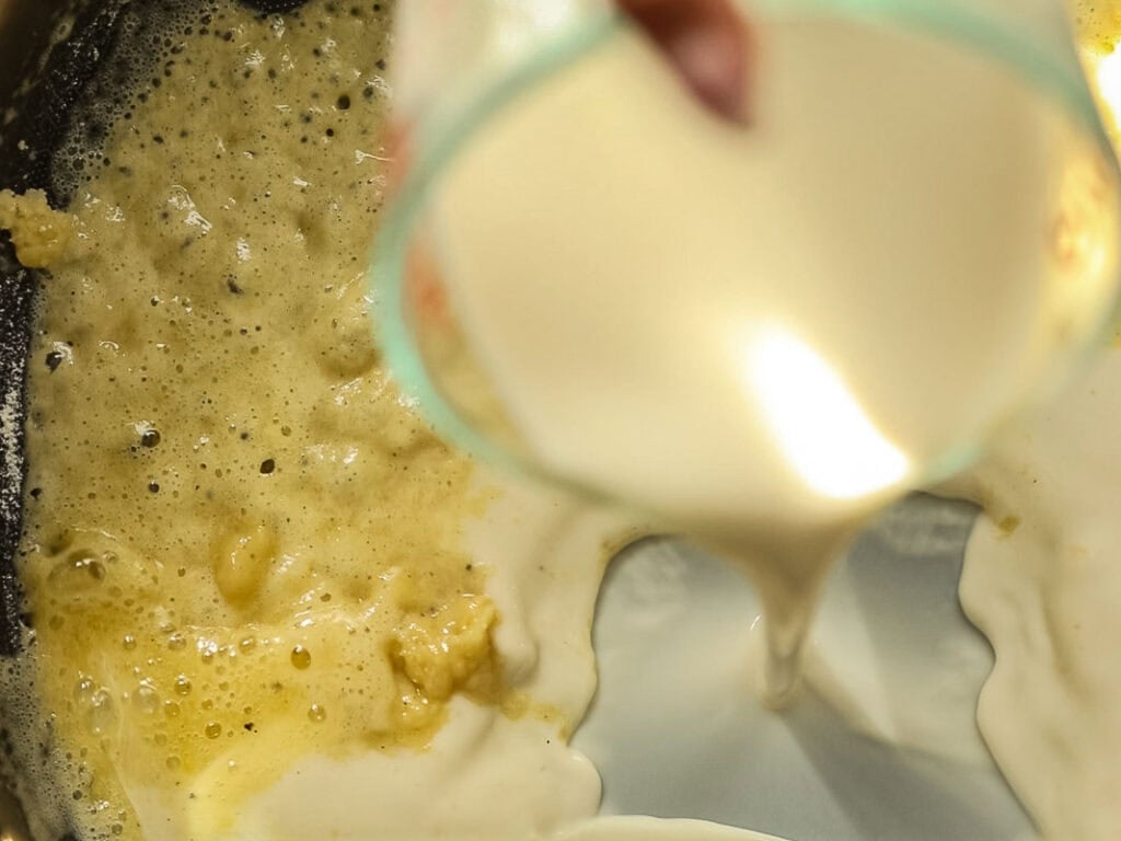 Close-up of a creamy liquid, likely milk or cream, being poured into a pan with bubbling melted butter and flour, suggesting the start of making a sauce or roux.