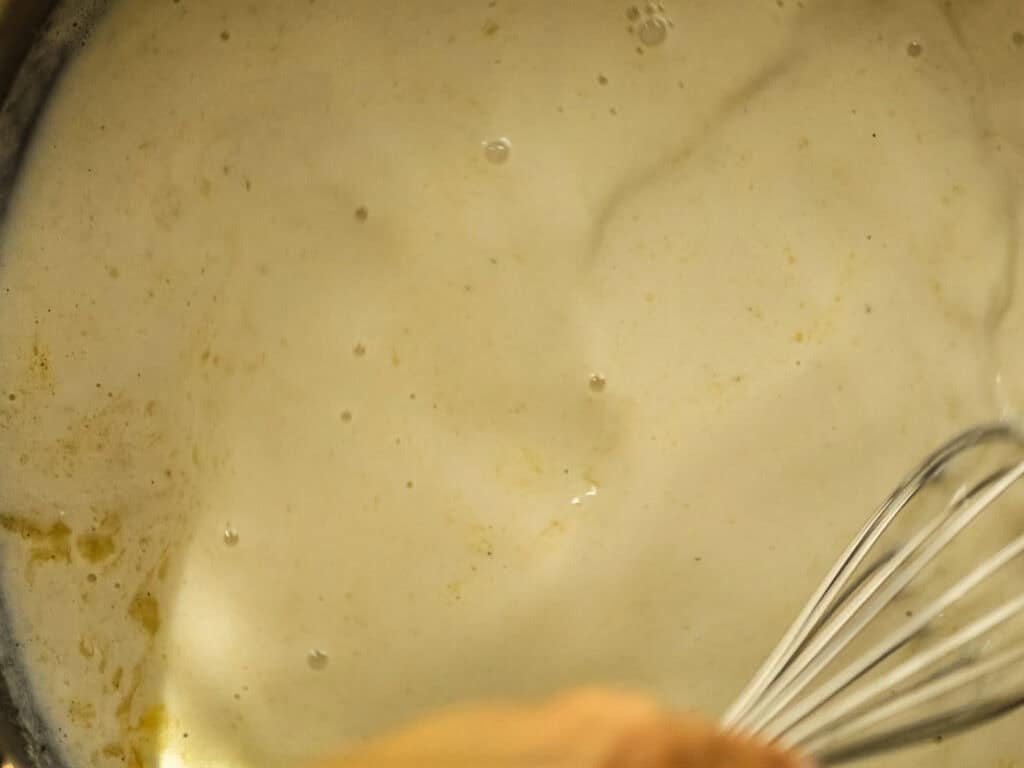 A close-up of a whisk stirring a creamy, foamy liquid in a pot, with some bubbles and a light yellow tint visible on the surface.