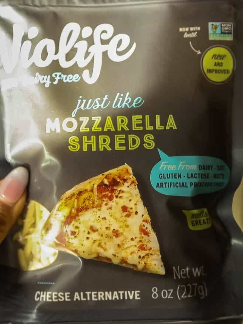 A hand holds a pack of Violife Just Like Mozzarella Shreds, a dairy-free cheese alternative. The packaging shows a slice of pizza and highlights that it is free from dairy, gluten, lactose, soy, and preservatives.