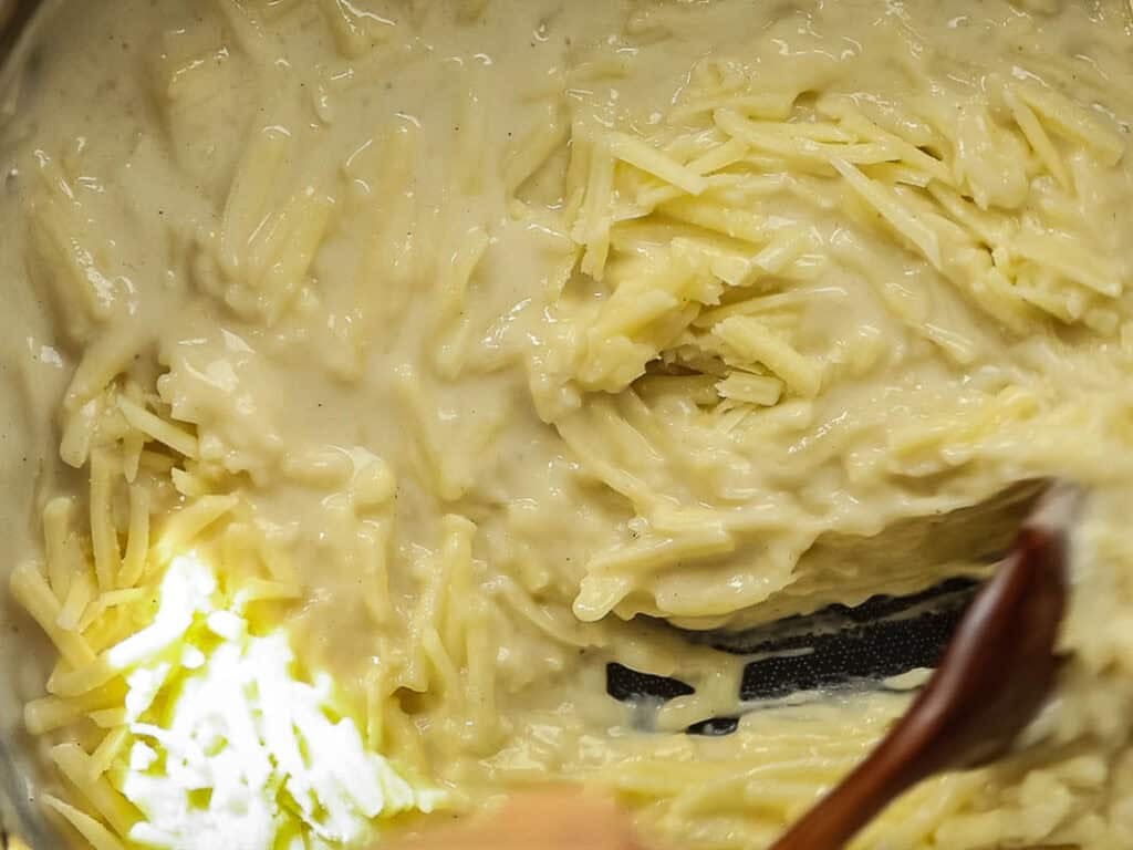 Close-up of shredded cheese being stirred into a creamy white sauce with a wooden spoon.