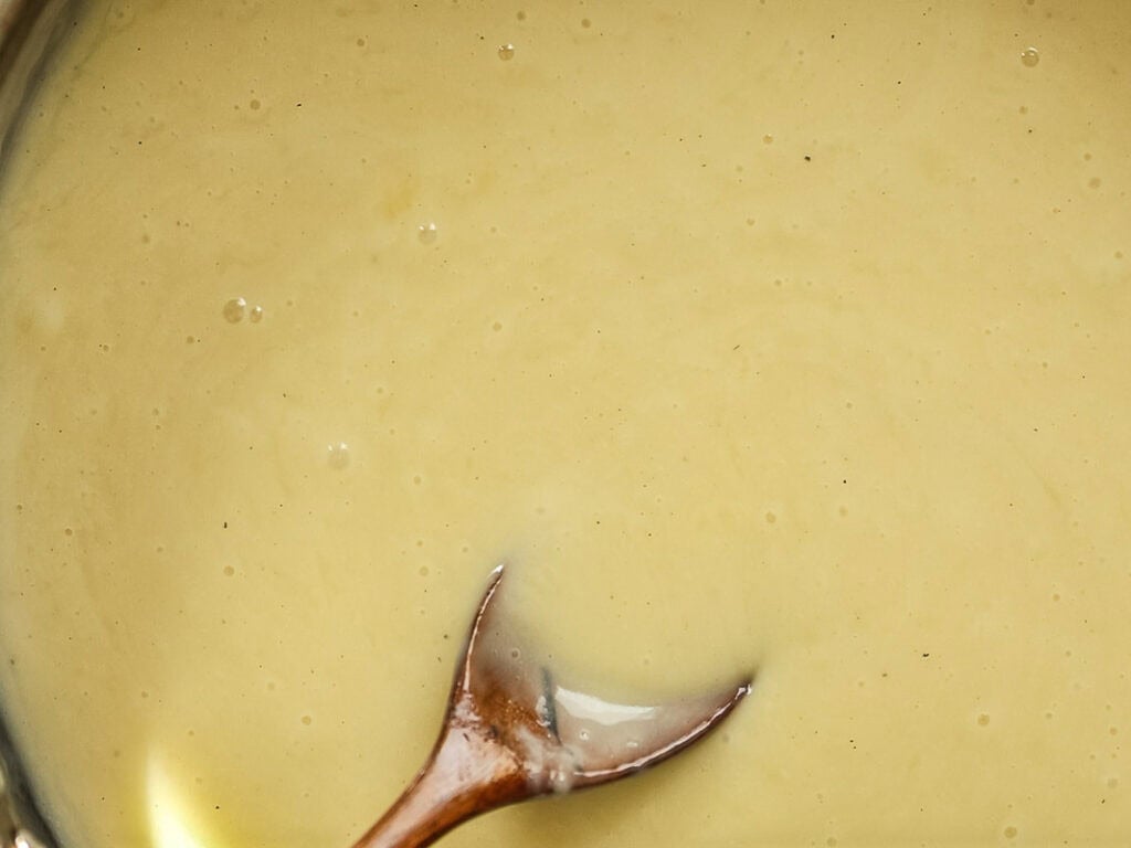 A close-up of a creamy, light yellow sauce being stirred with a wooden spoon. The texture appears smooth with a few small bubbles on the surface.