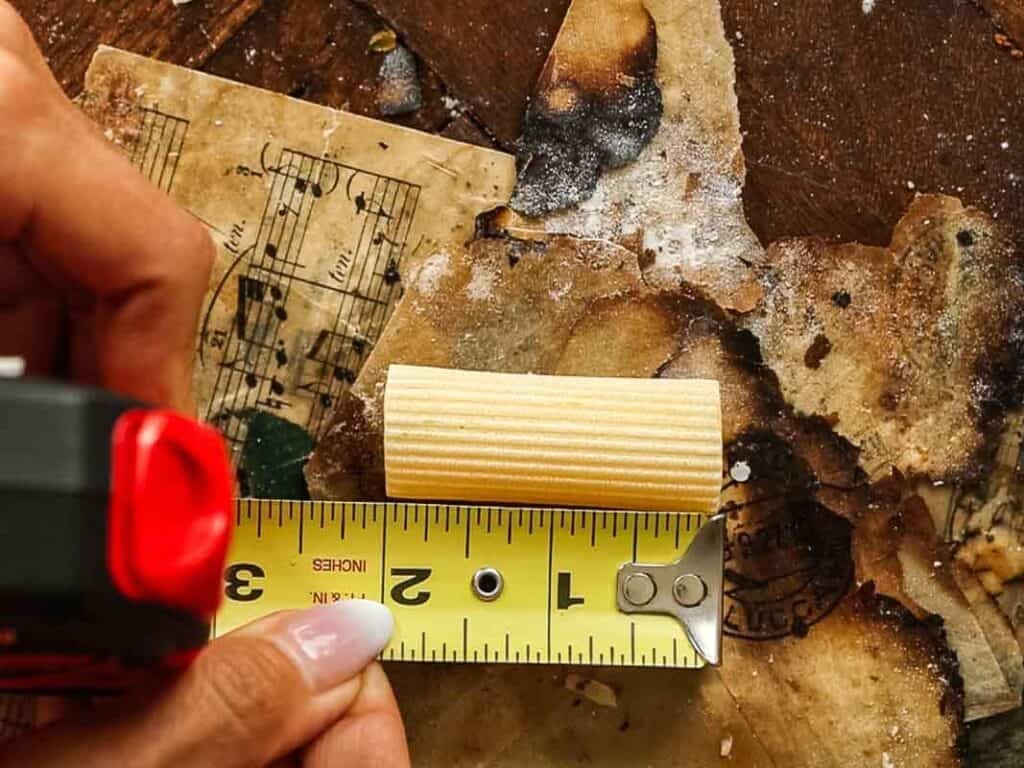 A hand holds a tape measure against a small wooden dowel on a cluttered, stained surface with old, burnt paper and sheet music in the background.