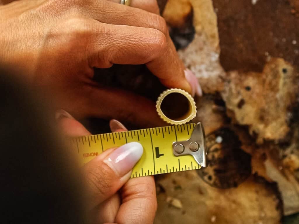 A person measures the diameter of a round pasta piece with a yellow tape measure on a rustic, brown surface.