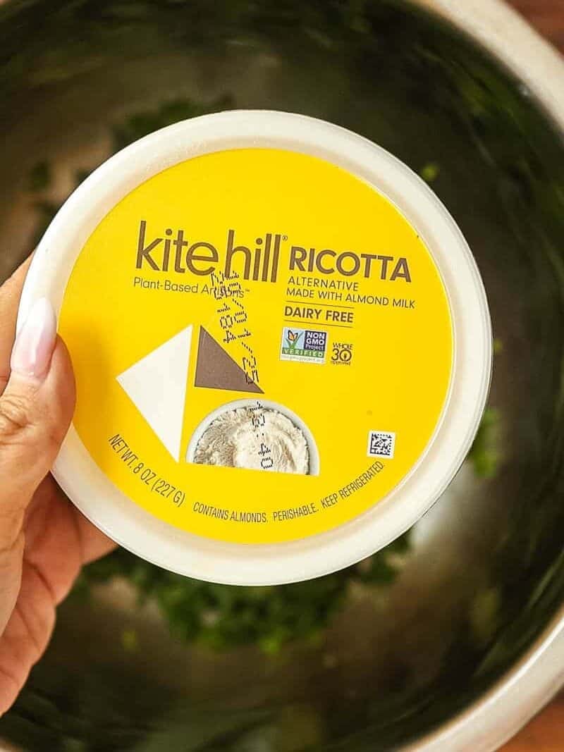 A hand holds a container of Kite Hill Ricotta, a plant-based, dairy-free cheese made from almond milk, over a metal bowl with chopped green herbs inside.