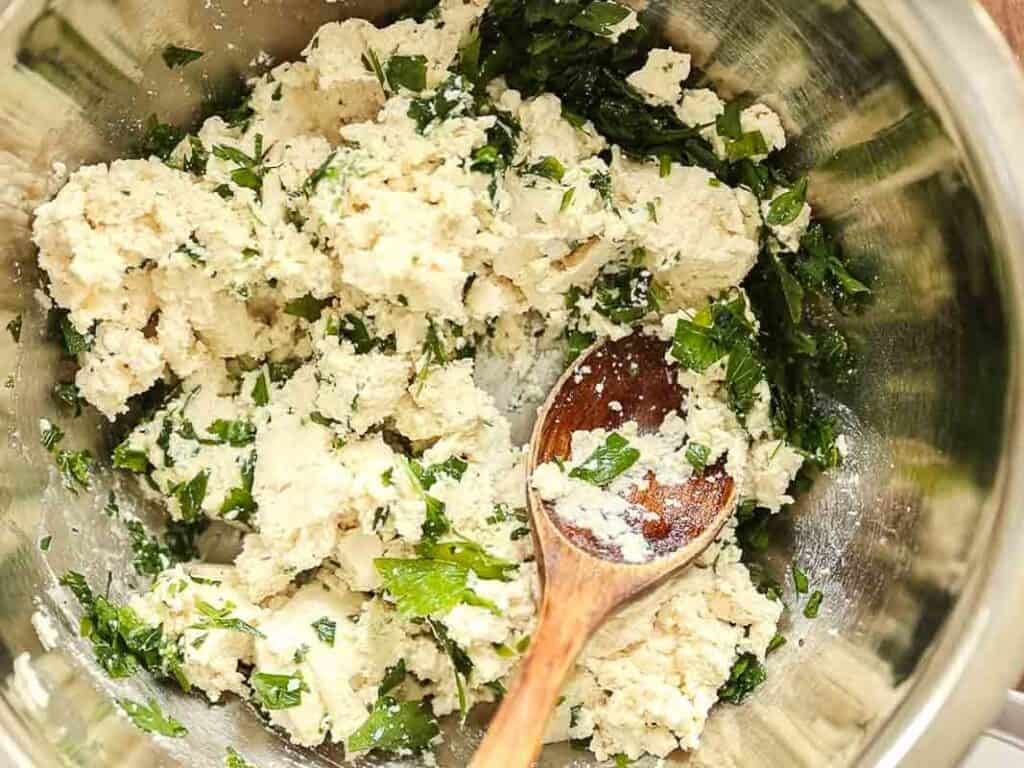 A metal mixing bowl filled with crumbled ricotta cheese mixed with chopped fresh herbs, being stirred with a wooden spoon.