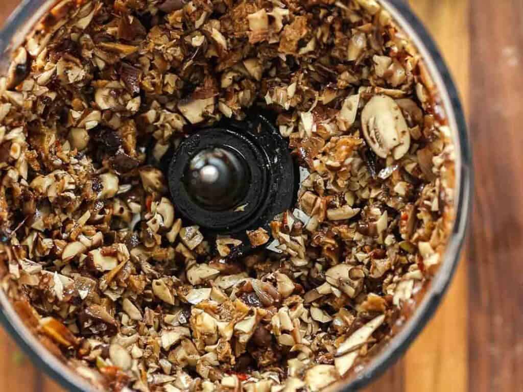 Chopped mushrooms and other ingredients inside a food processor, viewed from above, with the blades in the center, ready to be blended further.