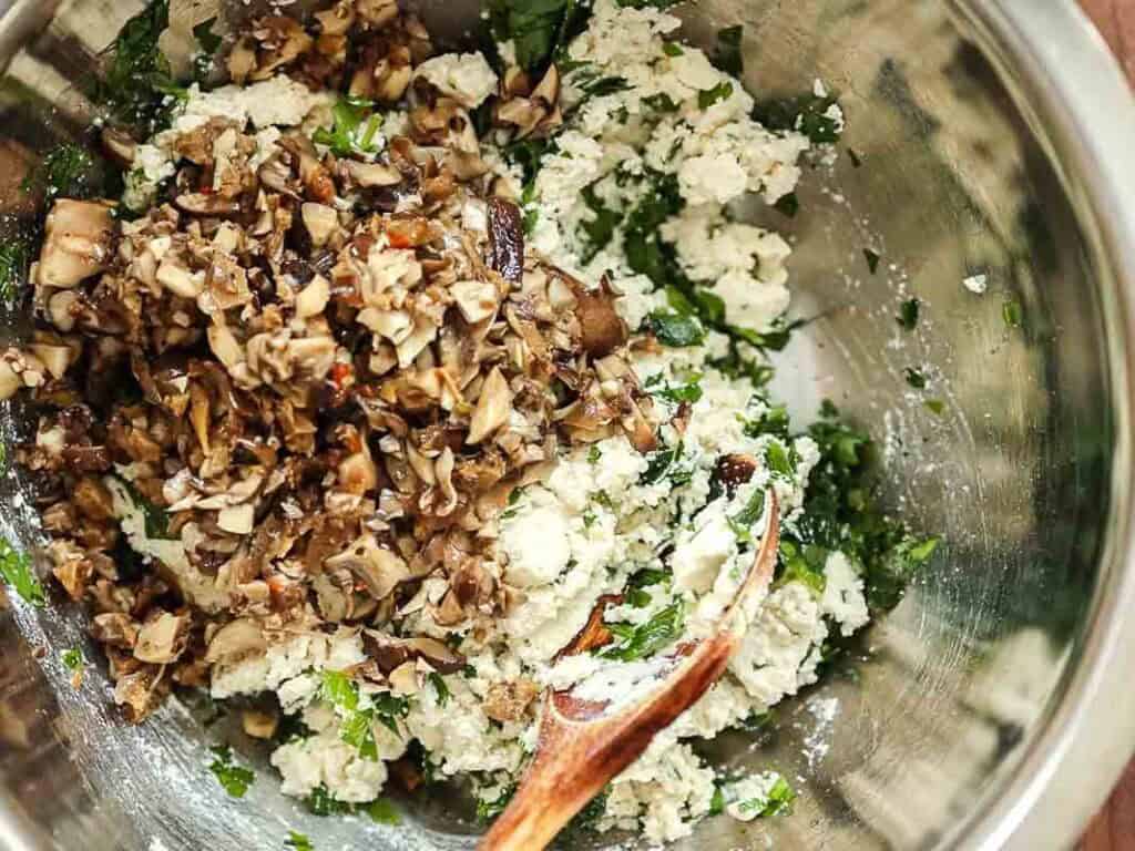 A metal bowl contains a mixture of chopped mushrooms, ricotta, and fresh herbs being mixed with a wooden spoon.