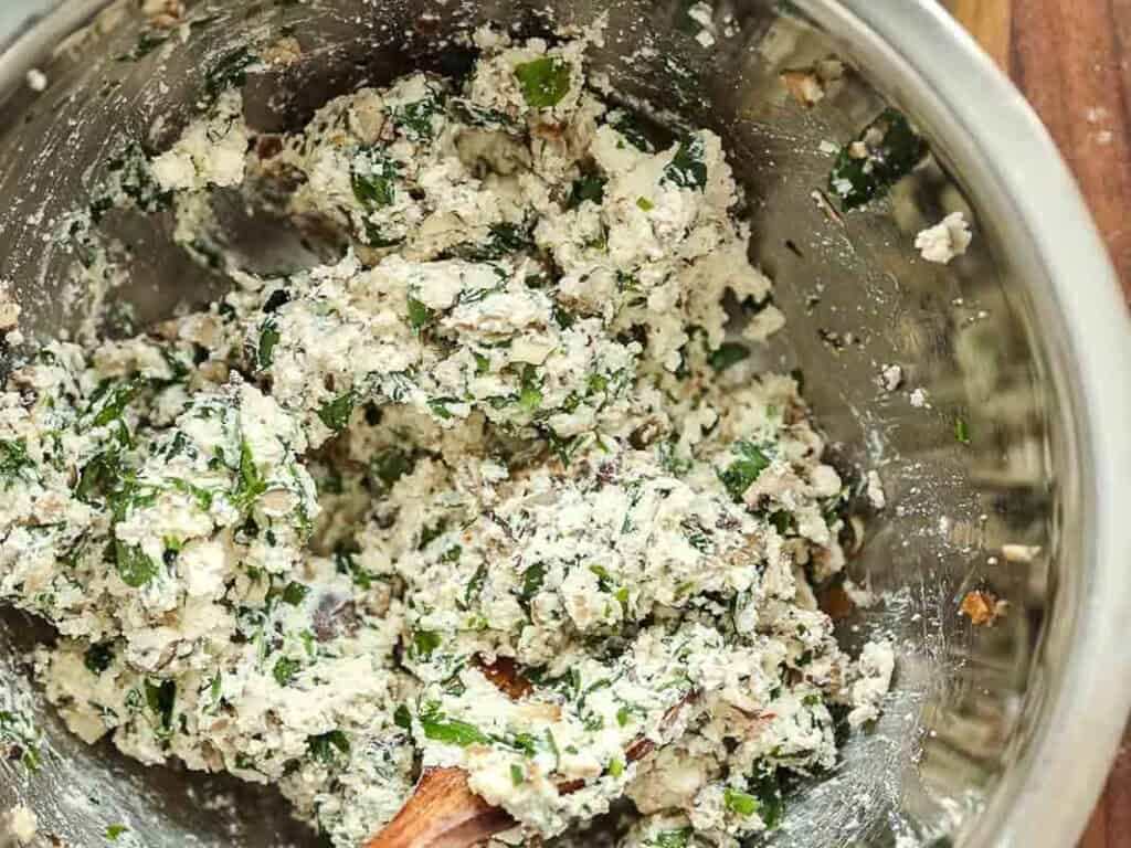 A metal mixing bowl containing a mixture of crumbled cheese, chopped greens, and herbs being combined with a wooden spoon.