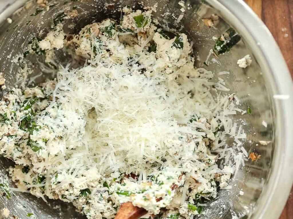 A metal mixing bowl filled with a mixture of ricotta cheese, herbs, and shredded parmesan cheese. A wooden spoon is partially visible in the bowl.