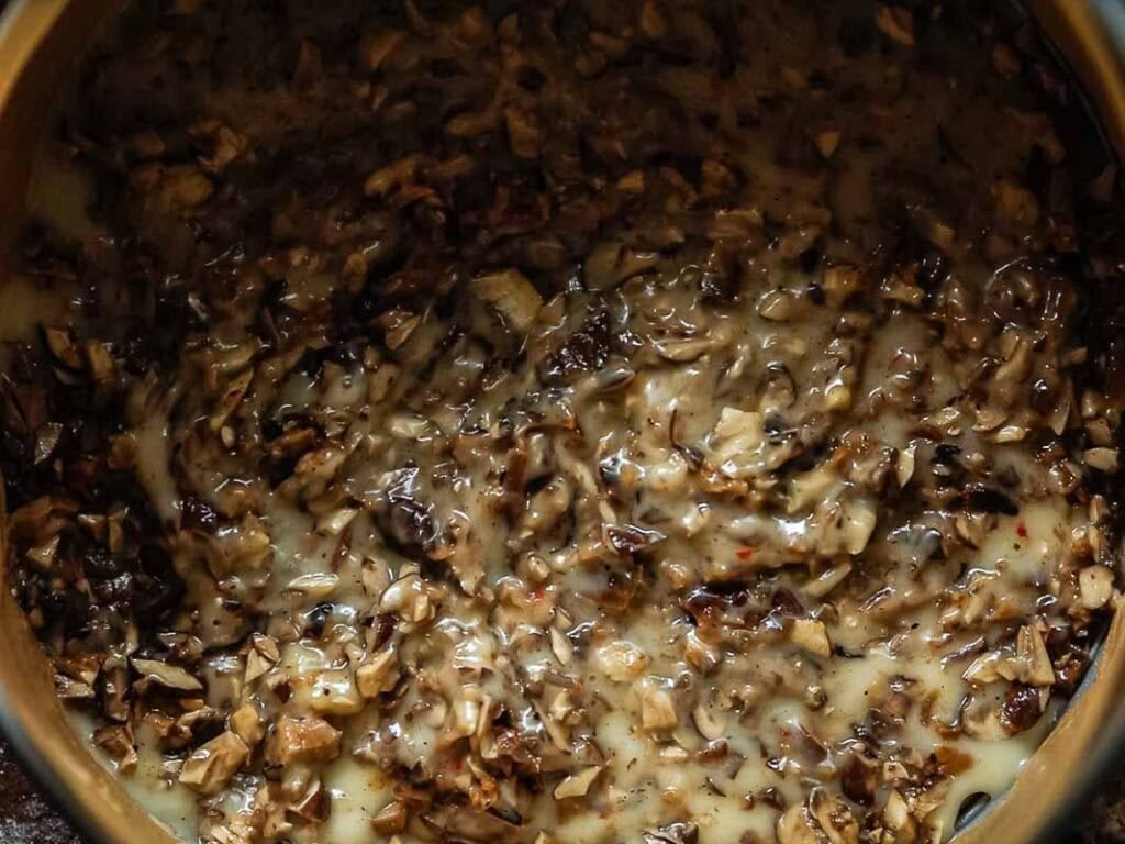 A close-up of a creamy mixture with finely chopped mushrooms in a pan, showing a thick consistency and a blend of brown and beige tones.