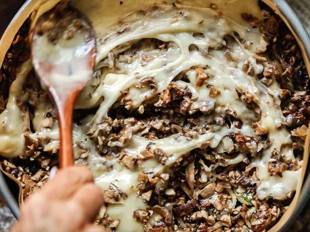 a hand holding wooden spoon mixes béchamel with minced mushroom mixture.