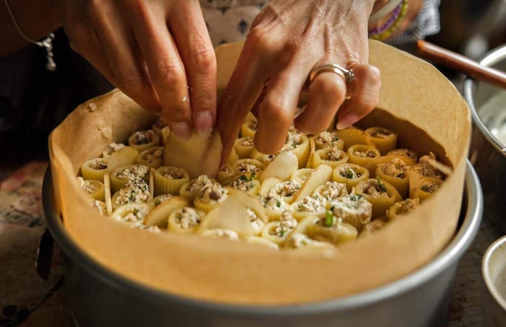 someone's hands adding sliced potatoes to rigatoni stuffed with ricotta in a springform pan lined with parchment paper.