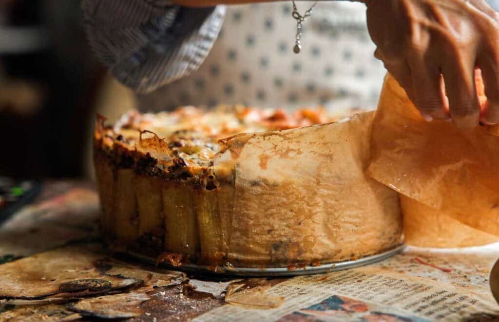 someone peeling back the parchment paper off the side of mushroom rigatoni pie.